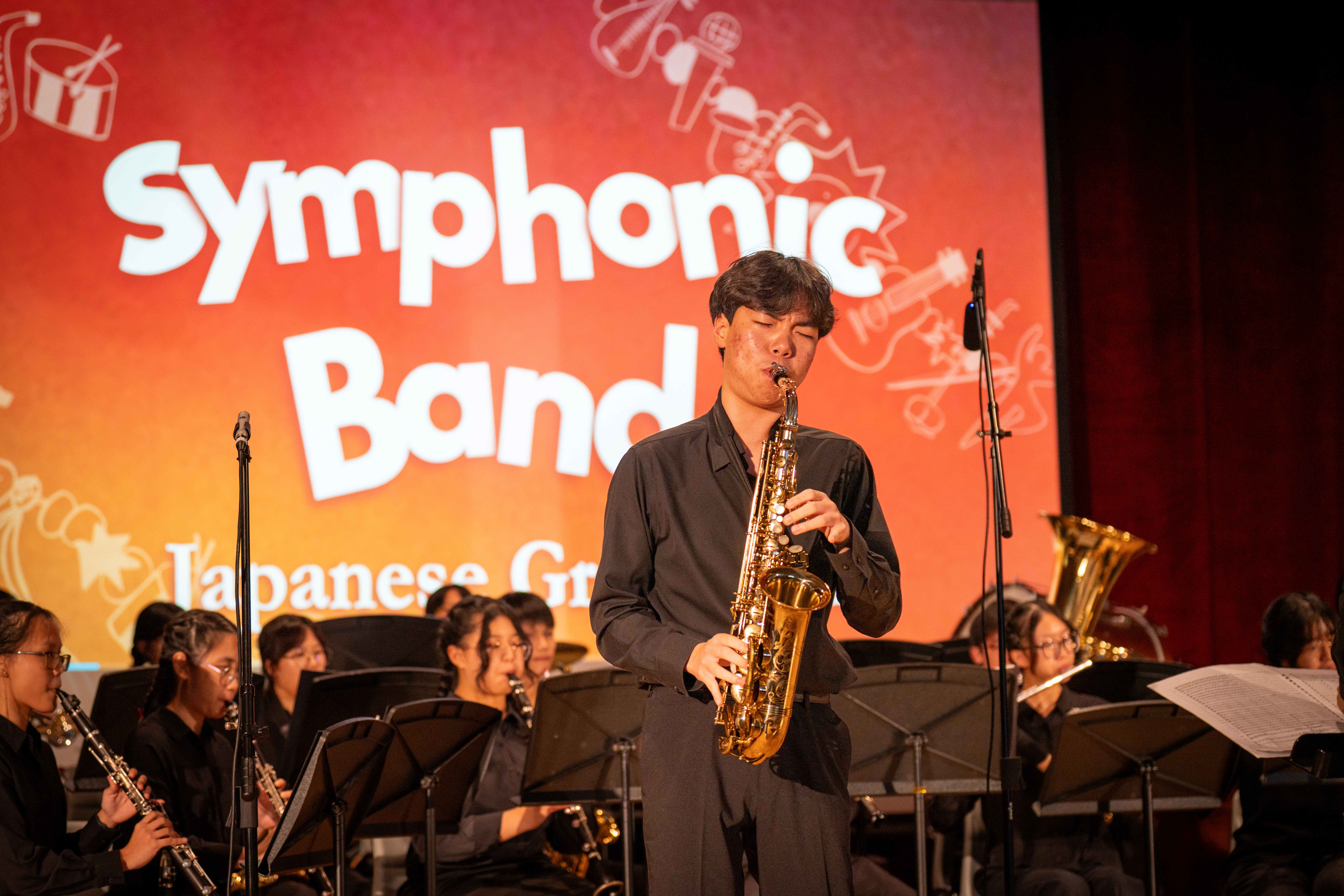 A young male student in a black outfit performs a saxophone solo on stage in front of the TMJC Symphonic Band, with a red-orange "Symphonic Band" backdrop behind him and fellow band members seated and playing in the background.